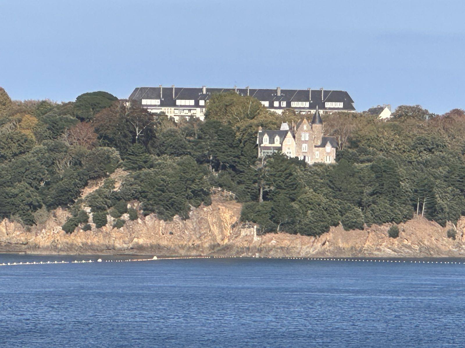 Vue prise depuis la rive d'en face : sur la hauteur boisée, un long immeuble d'ardoise domine une villa à tourelle ; en contrebas, les rochers, une ligne de bouées jaunes et la mer.