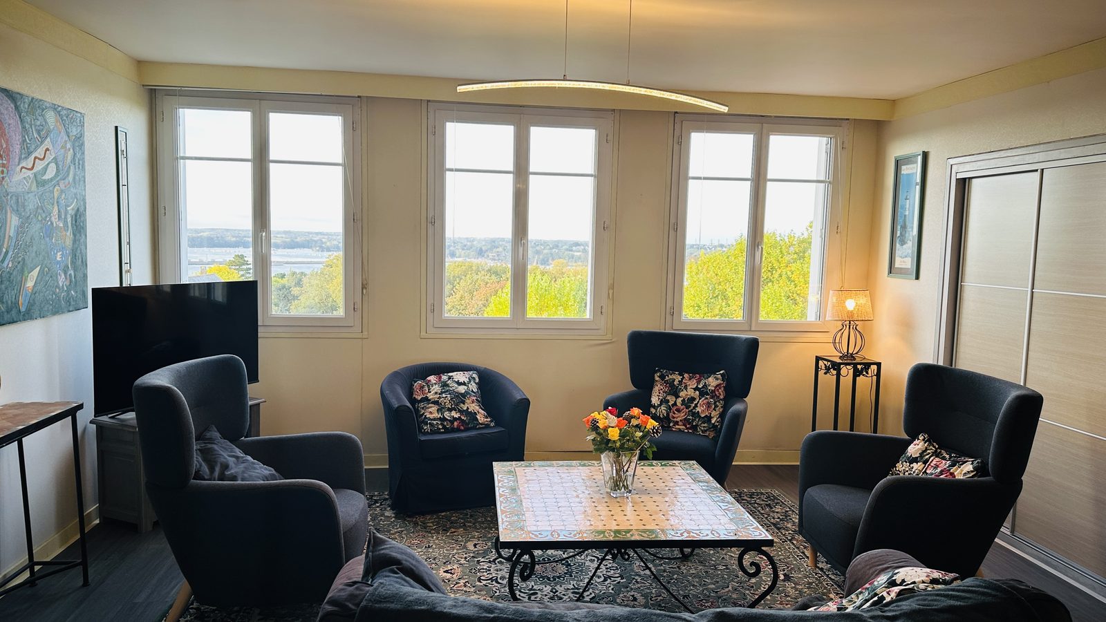 Le séjour au sixième étage : quatre fauteuils gris aux coussins fleuris et un canapé autour d'une table basse à plateau de faïence ; trois fenêtres donnent sur la verdure et la mer.