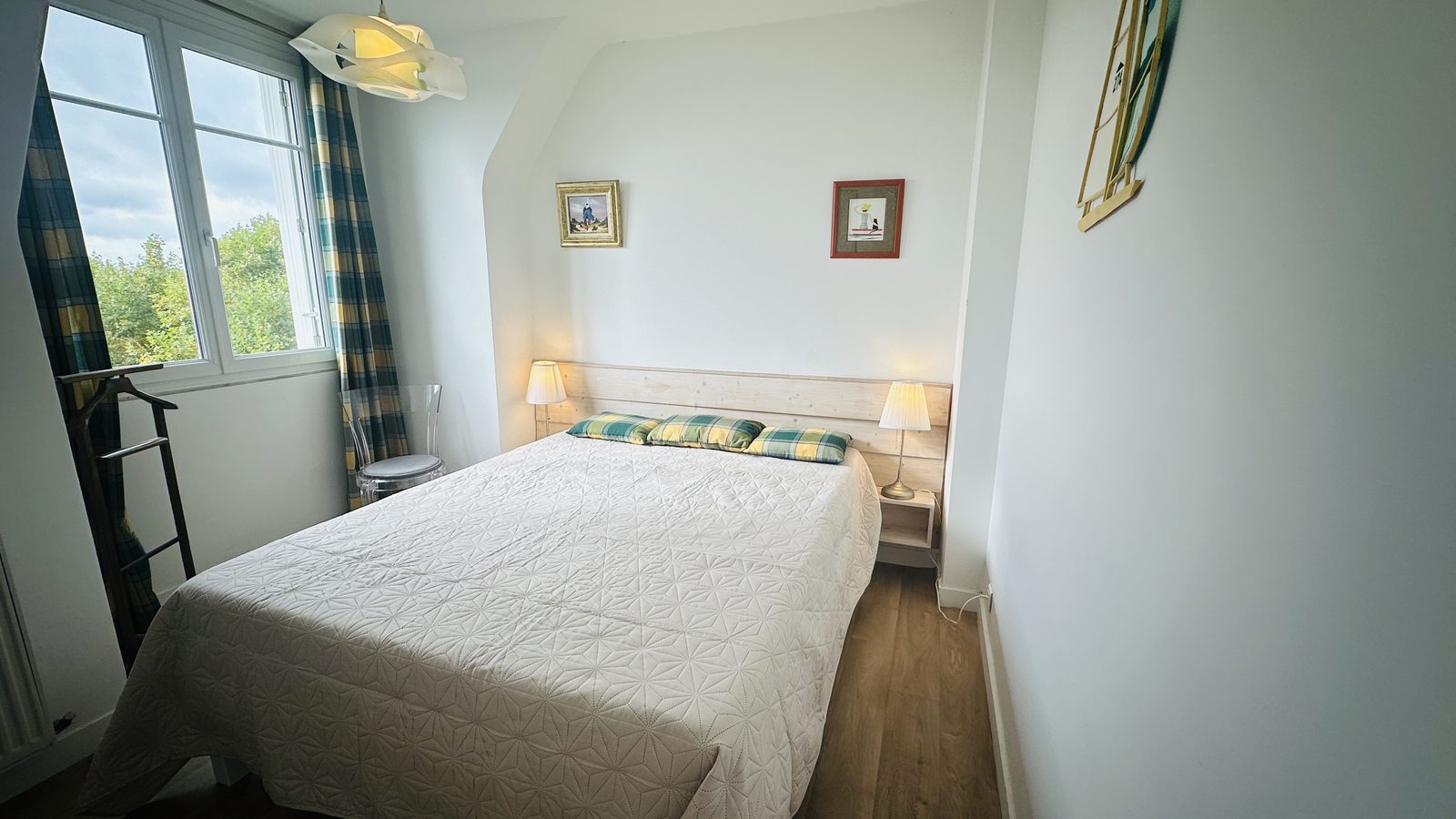 Chambre 1, sous rampant, murs blancs : le grand lit au couvre-lit blanc et ses coussins écossais, tête de lit en bois clair, deux tables de chevet et leurs lampes ; à gauche, un valet de chambre et la fenêtre aux rideaux écossais ouverte sur la verdure.