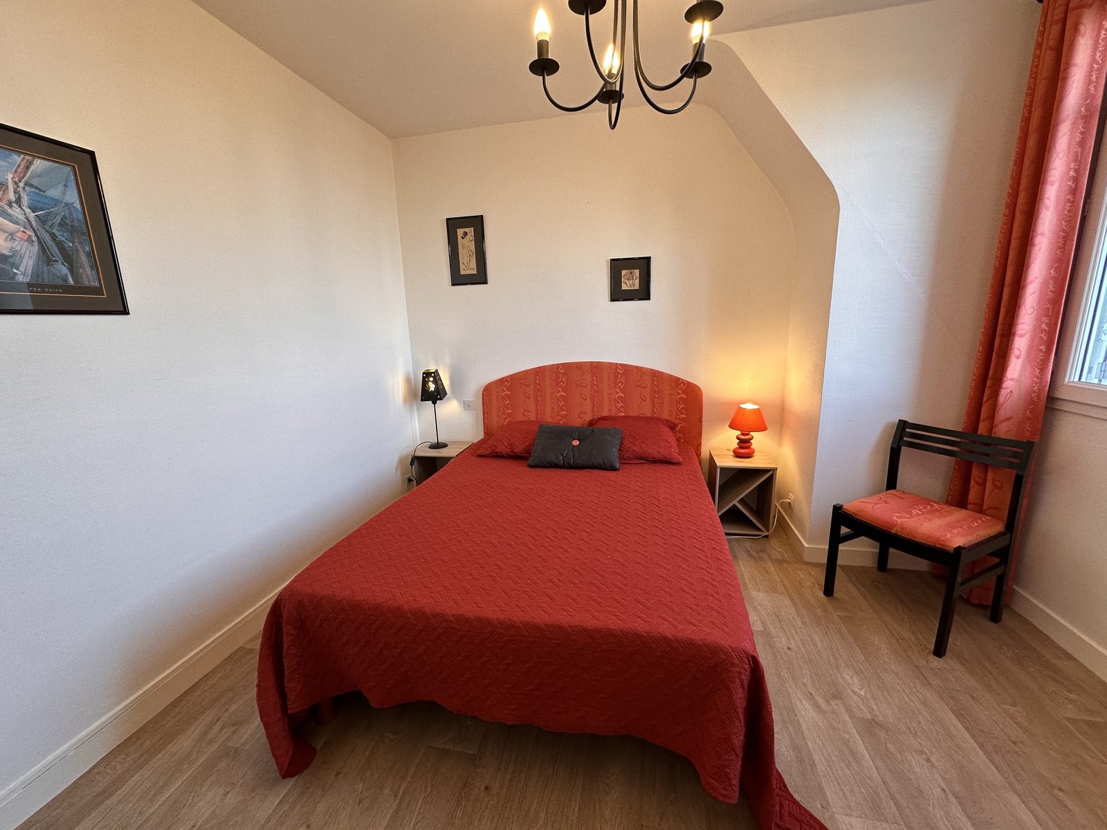 Chambre sous rampant : lit double au couvre-lit rouge, lustre à bougies, deux tables de chevet avec lampes, une chaise. Sol en lames de bois clair.