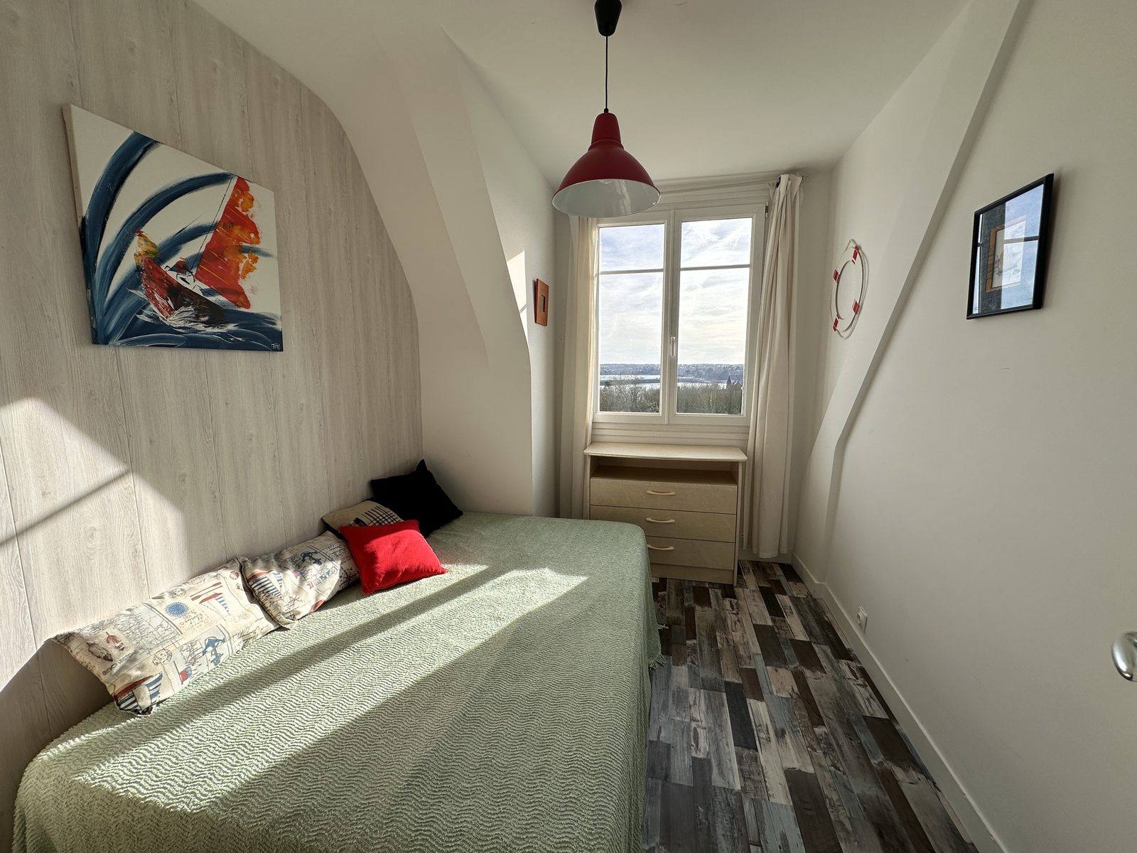 Chambre mansardée : un lit double au couvre-lit vert clair, garni de coussins et poussé contre le mur, une suspension rouge, une commode, et une fenêtre par laquelle on aperçoit la mer. Au mur, une toile de voilier.
