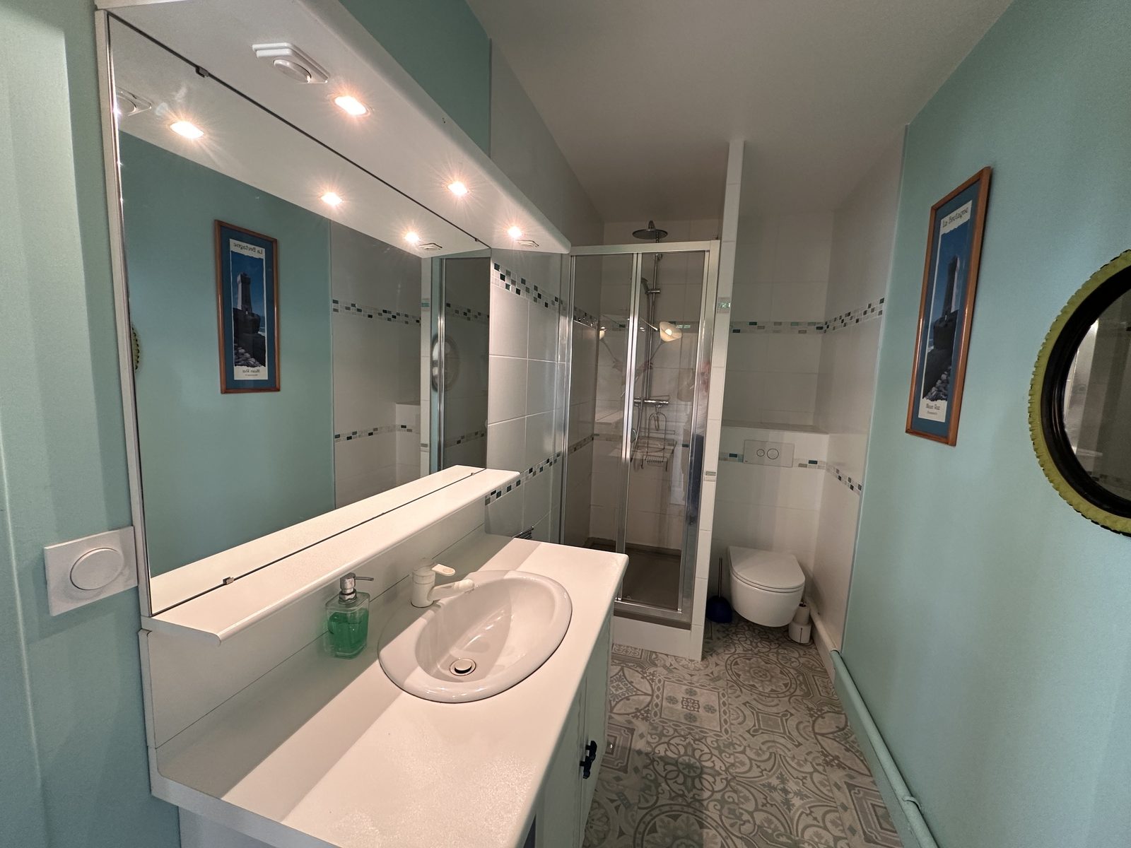 Seconde salle d'eau, aux murs vert d'eau : un long miroir éclairé de spots au-dessus d'une vasque, une cabine de douche vitrée, un wc suspendu, et un sol à motifs de carreaux de ciment.