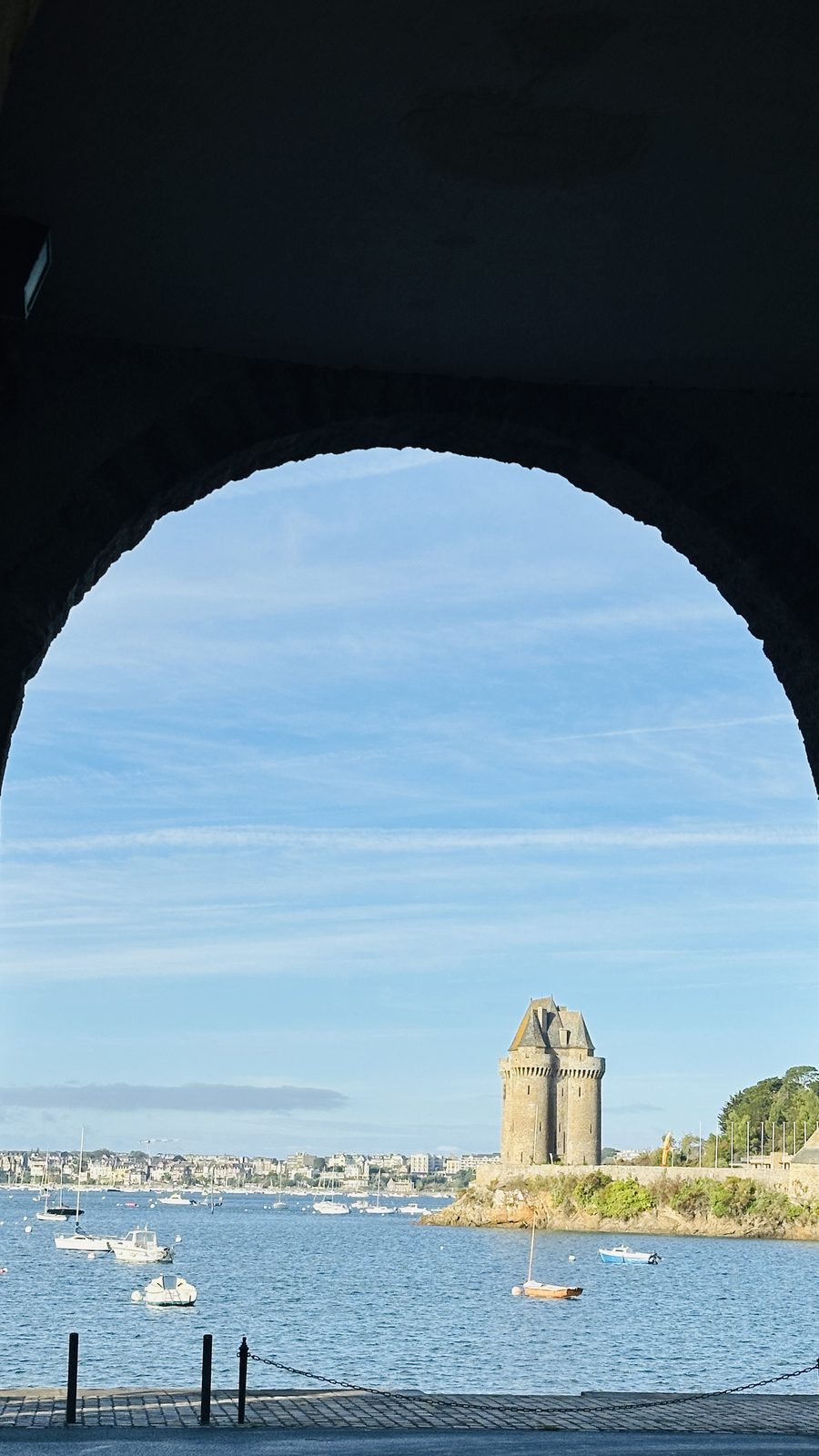 La Tour Solidor vue à travers une arche de pierre du quai : le donjon de granit à trois tours sur son rocher, des bateaux au mouillage et Dinard au loin.